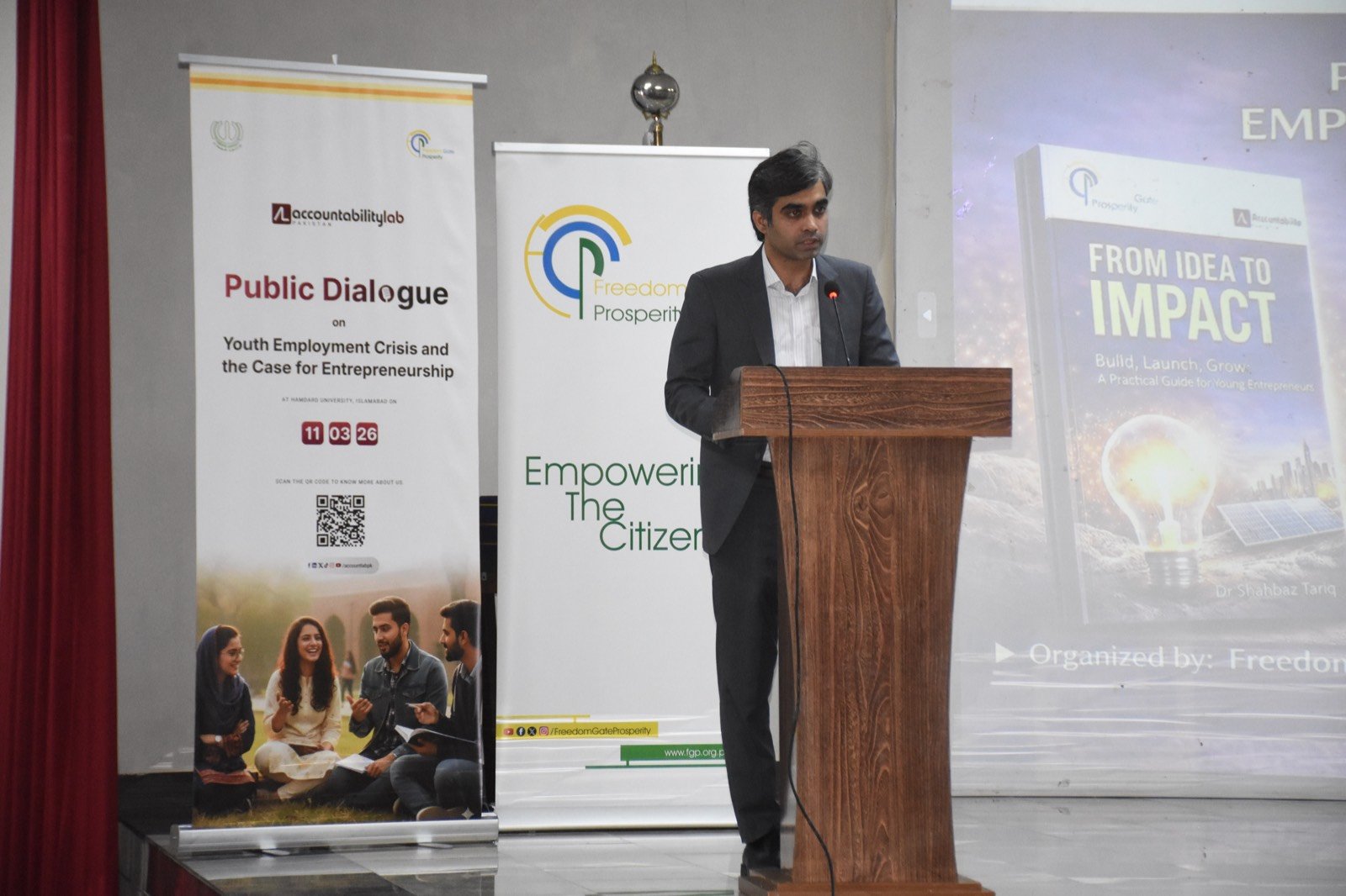 A speaker addressing the public dialogue with the From Idea to Impact book featured on screen
